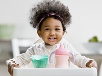 A toddler smiling in a highchair with a container of cereal and a sippy cup.